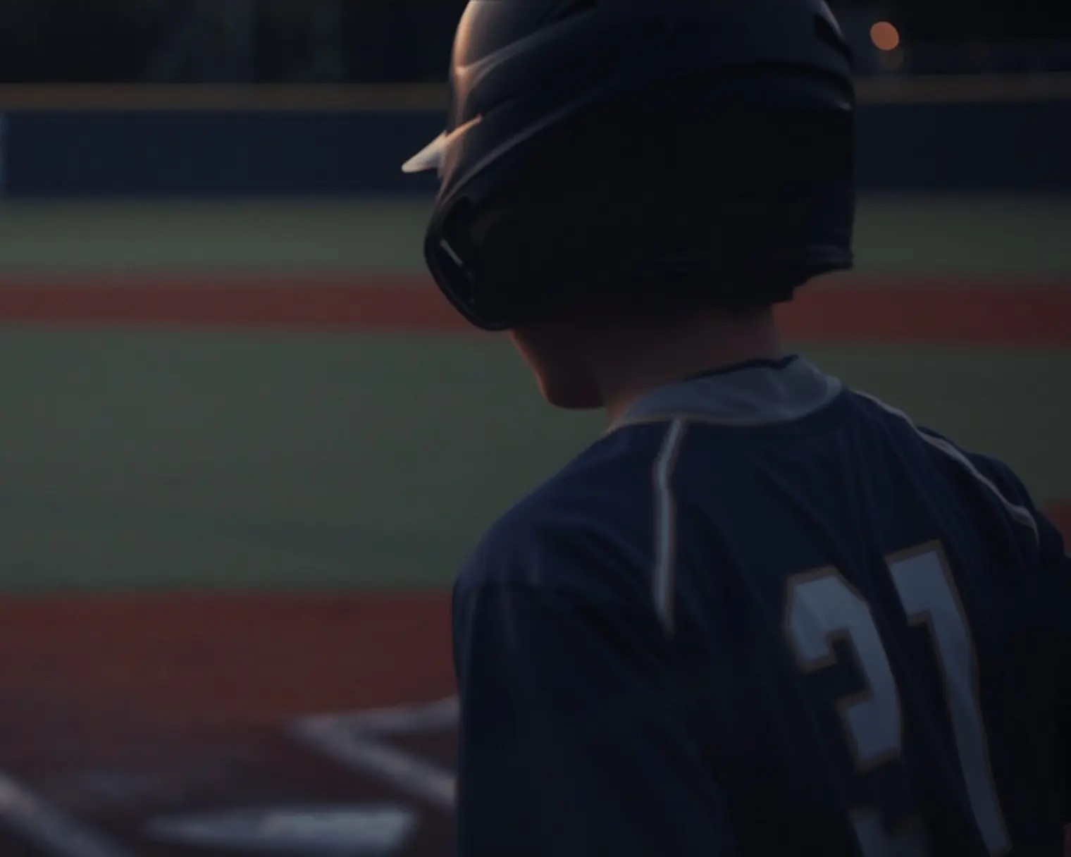 Youth batter at home plate at dusk, viewed from behind